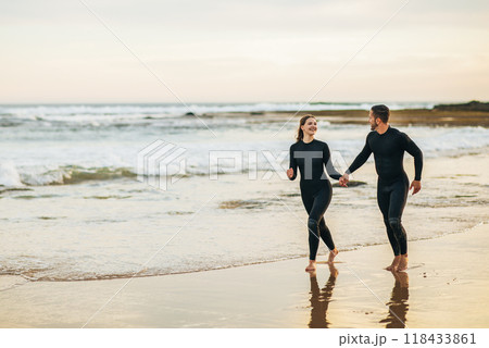 A happy couple runs along the sandy beach holding hands, both wearing black wetsuits. The ocean waves gently lap at the shore behind them as the sun sets in the distance, copy space 118433861