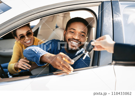 This image depicts a happy couple receiving the keys to their new car from a dealership representative. The man is sitting in the drivers seat of a white sedan and is reaching out to take the keys 118433862