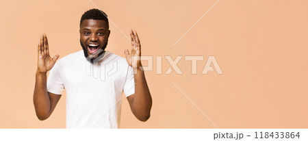 A young black man wearing a white t-shirt smiles and looks shocked while raising his hands in the air. He has a light brown complexion and a full beard. He is standing in front of a peach background. 118433864