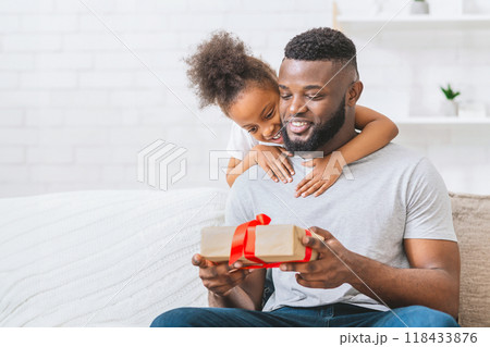 Black father sits on a couch with his daughter embracing him from behind. The father is opening a gift wrapped in brown paper and red ribbon. Both appear to be smiling and happy. Black father sits on a couch with his daughter embracing him from behind. The father is opening a gift wrapped in brown paper and red ribbon. Both appear to be smiling and happy. 118433876