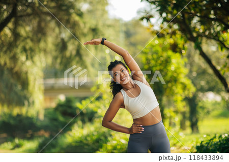 A woman in a white tank top and gray leggings stretches her arm overhead while smiling. She is in a park with lush green trees and foliage, and the sun is shining brightly. 118433894