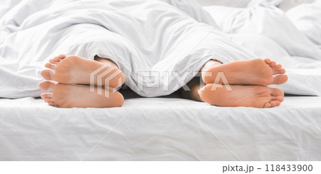 This image shows two pairs of feet sticking out from under white sheets in a bed. The feet are bare and the sheets are white and wrinkle-free. 118433900
