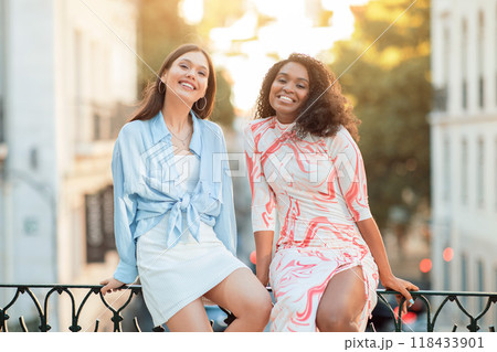Two multiethnic young women sit on a railing in a city setting, smiling and laughing, enjoying each others company. The photo captures a moment of joy and friendship Two multiethnic young women sit on a railing in a city setting, smiling and laughing, enjoying each others company. The photo captures a moment of joy and friendship 118433901