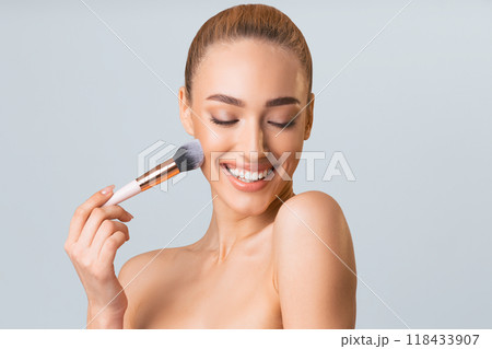 A young woman with light brown hair pulled back into a bun smiles as she applies makeup with a blush brush. She is looking down at the brush with her eyes closed and her mouth slightly open. A young woman with light brown hair pulled back into a bun smiles as she applies makeup with a blush brush. She is looking down at the brush with her eyes closed and her mouth slightly open. 118433907