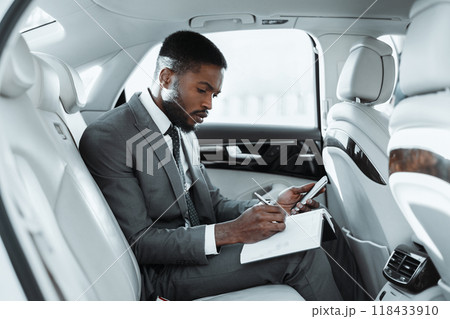 African American businessman sits in the backseat of a car, working on a notepad and using a smartphone. He is dressed in a suit and tie, and appears to be focused on his task. African American businessman sits in the backseat of a car, working on a notepad and using a smartphone. He is dressed in a suit and tie, and appears to be focused on his task. 118433910