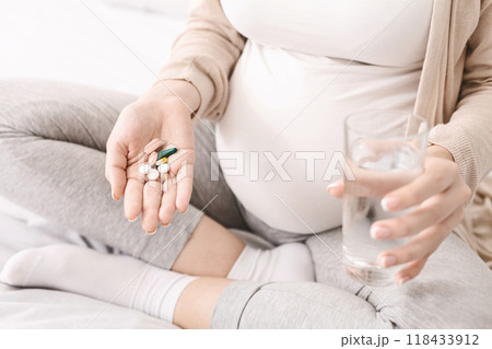 A pregnant woman is seen holding a pill in one hand and a glass of water in the other, preparing to take medication. The image shows the importance of taking prescribed medication during pregnancy A pregnant woman is seen holding a pill in one hand and a glass of water in the other, preparing to take medication. The image shows the importance of taking prescribed medication during pregnancy 118433912