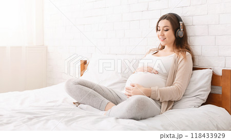 Young millennial pregnant woman listening to music through wireless headphones, relaxing on bed at home, panorama with free space 118433929