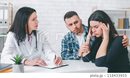 A doctor in a white coat sits at a desk with a clipboard and pen, listening attentively as a young couple shares their concerns. The woman is visibly upset, holding her head in her hands A doctor in a white coat sits at a desk with a clipboard and pen, listening attentively as a young couple shares their concerns. The woman is visibly upset, holding her head in her hands 118433933