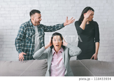 A woman sits on a couch with her hands over her ears as a man yells at another woman standing behind the couch. Woman standing behind the couch with her hand on her forehead. A woman sits on a couch with her hands over her ears as a man yells at another woman standing behind the couch. Woman standing behind the couch with her hand on her forehead. 118433934