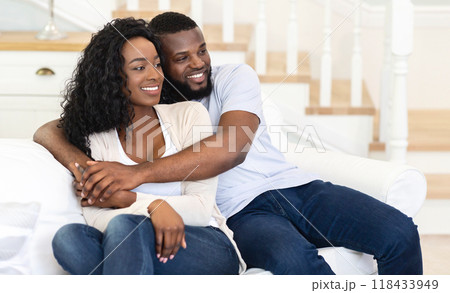 A man and woman are sitting on a white couch in a home. The man has his arm around the woman and they are both looking to the side. 118433949