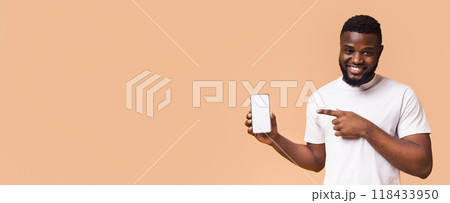 A young black man with a beard is smiling and holding a smartphone with a blank screen while pointing at it with his right hand, panorama with copy space 118433950