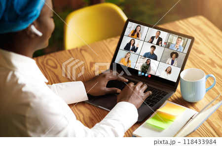 African American woman wearing a blue headband and white shirt sits at a wooden table, using a laptop to participate in a video conference meeting, cafe interior 118433984