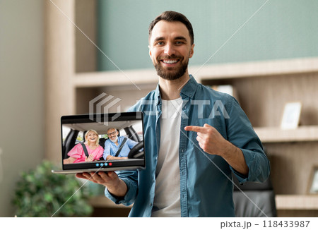 A man stands in a home office, smiling and pointing at his laptop screen, which shows a video call with an elderly couple sitting in a car. A man stands in a home office, smiling and pointing at his laptop screen, which shows a video call with an elderly couple sitting in a car. 118433987