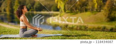 Side view of millennial woman in sports outfit meditating on yoga mat at lake in green park outside. Lady practicing breathing exercises in nature for inner peace and harmony outdoor, copy space Side view of millennial woman in sports outfit meditating on yoga mat at lake in green park outside. Lady practicing breathing exercises in nature for inner peace and harmony outdoor, copy space 118434087
