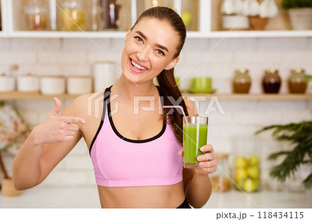 A woman in athletic wear smiles and holds a green smoothie in a glass with a straw. 118434115