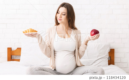 Healthy nutrition and pregnancy. Pregnant woman choosing between croissant and apple, sitting on bed at home Healthy nutrition and pregnancy. Pregnant woman choosing between croissant and apple, sitting on bed at home 118434120