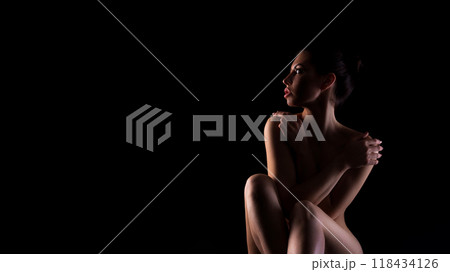 A woman sits in a dimly lit room, her body illuminated by a soft light source. She is facing away from the camera, with her arms wrapped around her body, copy space 118434126