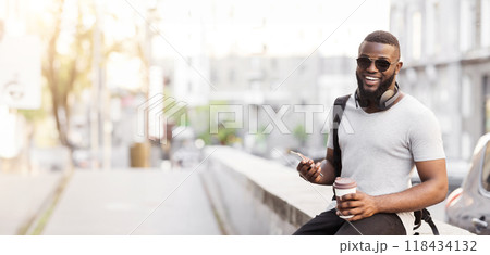 A man wearing sunglasses and a backpack sits on a ledge in an urban setting, holding a coffee cup and a smartphone. A man wearing sunglasses and a backpack sits on a ledge in an urban setting, holding a coffee cup and a smartphone. 118434132