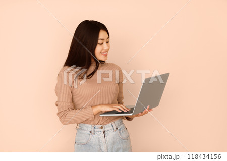 A young woman with long black hair smiles as she uses a laptop against a peach-colored background. A young woman with long black hair smiles as she uses a laptop against a peach-colored background. 118434156