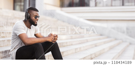 A man wearing headphones and sunglasses sits on a set of stairs, listening to music. 118434189