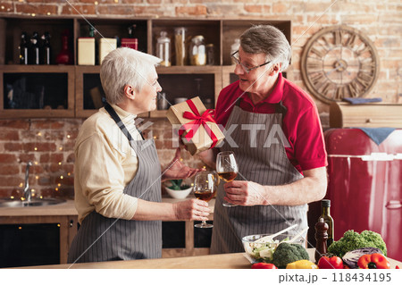 A senior couple celebrates with a bottle of wine and a gift, smiling at each other in a kitchen. The man is holding the gift, and the woman is holding a glass of wine. 118434195