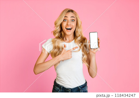 This image shows a young woman with long, blonde hair wearing a white t-shirt and blue jeans standing in front of a pink background. She is holding a smartphone with a blank screen This image shows a young woman with long, blonde hair wearing a white t-shirt and blue jeans standing in front of a pink background. She is holding a smartphone with a blank screen 118434247