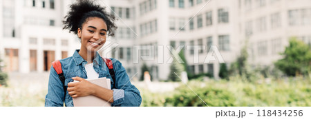 Portrait Of Successful Student. Happy African American Teenager Girl Posing Holding Books Smiling Looking At Camera Standing Near Modern University Building. Modern Education, Copy Space Portrait Of Successful Student. Happy African American Teenager Girl Posing Holding Books Smiling Looking At Camera Standing Near Modern University Building. Modern Education, Copy Space 118434256