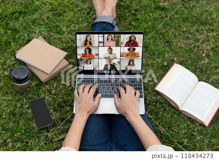 Lady is sitting on green grass, using a laptop for a video call. There are multiple people on the screen, and the person is typing on the keyboard. 118434273
