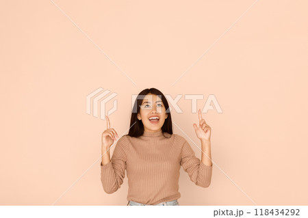 A woman in a brown sweater points both index fingers upward against a beige background. 118434292