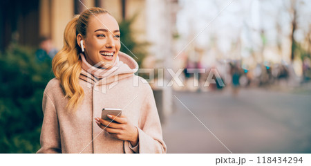 A young woman with long blonde hair walks down a city street, smiling and looking to the side. She is wearing a pink hoodie and coat and holding a phone in one hand, panorama with copy space 118434294