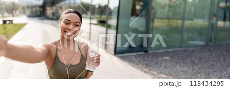 African American woman taking selfie with cellphone, recording video for her blog, holding water bottle on street. Black lady influencer communicating to followers, copy space African American woman taking selfie with cellphone, recording video for her blog, holding water bottle on street. Black lady influencer communicating to followers, copy space 118434295