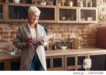 A senior woman stands in her kitchen, wearing a gray cardigan and jeans. She holds a red mug in one hand and reads a newspaper with the other, copy space 118434390