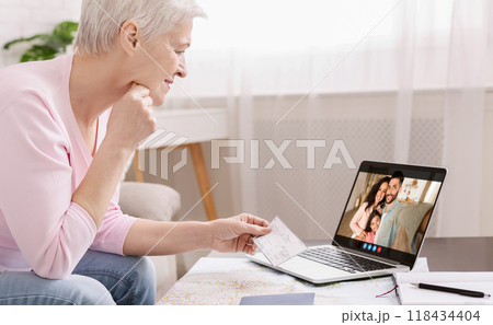 A senior woman is sitting at a table with a laptop in front of her. She is smiling and holding up a check for her family on the video call. The family is smiling back at her. A senior woman is sitting at a table with a laptop in front of her. She is smiling and holding up a check for her family on the video call. The family is smiling back at her. 118434404