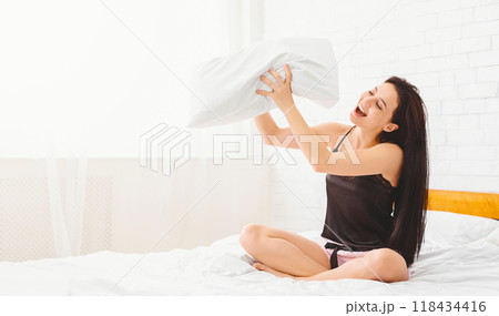 A woman is seen seated on a bed, clutching a pillow in her arms, singing. The room is minimally decorated, with only a bedside table visible in the background, copy space A woman is seen seated on a bed, clutching a pillow in her arms, singing. The room is minimally decorated, with only a bedside table visible in the background, copy space 118434416