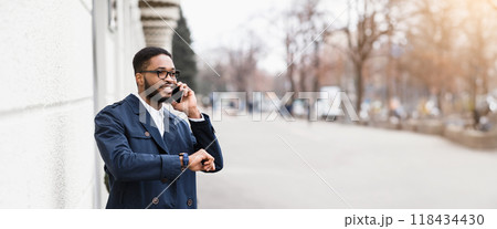 Black businessman in formal outfit have phone conversation outdoors, checking time, standing by modern building on the street, panorama with copy space 118434430