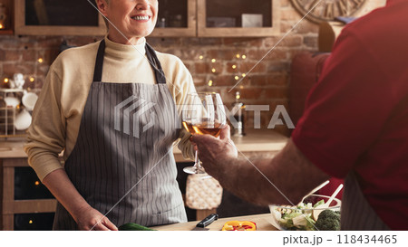 A senior woman in an apron stands in a kitchen and smiles as her partner toasts her with a glass of wine. The kitchen is decorated with exposed brick and a farmhouse style, cropped A senior woman in an apron stands in a kitchen and smiles as her partner toasts her with a glass of wine. The kitchen is decorated with exposed brick and a farmhouse style, cropped 118434465