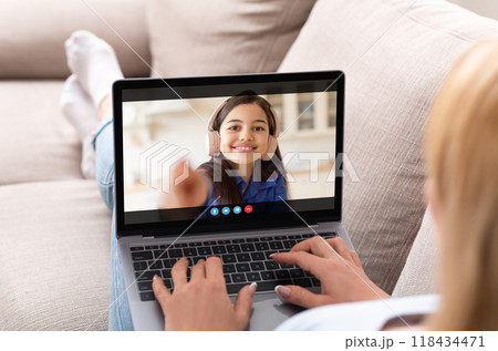 A woman is sitting on a couch, relaxing with her feet up, and chatting with a young girl on a laptop. The girl is smiling and waving to the camera, while the woman types on the keyboard. 118434471