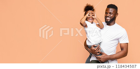 African American happy father and his little daughter smiling and playing together against a peach-colored backdrop. The father is holding his daughter in his arms, and she is playfully making glasses 118434507
