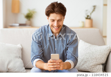 A man sits on a couch, smiling and looking at his phone. 118434515