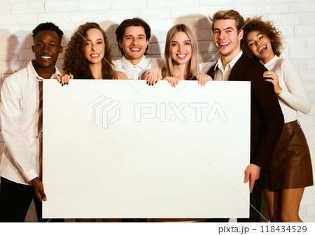 Cheerful Diverse Millennial Friends Holding Blank Board Standing Over White Brick Wall Indoor. Mockup Cheerful Diverse Millennial Friends Holding Blank Board Standing Over White Brick Wall Indoor. Mockup 118434529