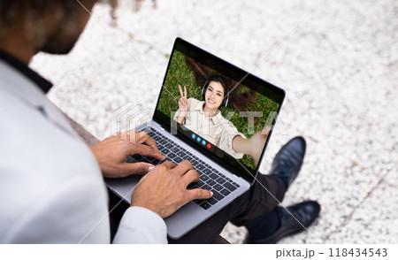 Black man sits outside on a bench, using a laptop to video chat with a woman. The woman is smiling and giving a peace sign. The man is typing on the laptop keyboard. 118434543