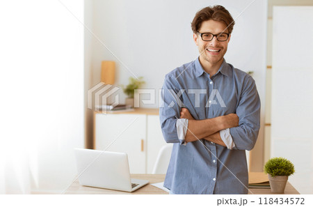A smiling man with crossed arms stands in a bright office setting. A smiling man with crossed arms stands in a bright office setting. 118434572