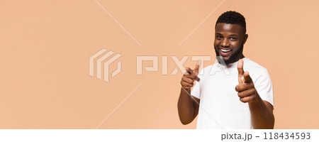 A smiling African American man is wearing a white T-shirt and pointing both index fingers at the camera. The background is a light peach color. This photo captures a moment of joy and confidence. A smiling African American man is wearing a white T-shirt and pointing both index fingers at the camera. The background is a light peach color. This photo captures a moment of joy and confidence. 118434593