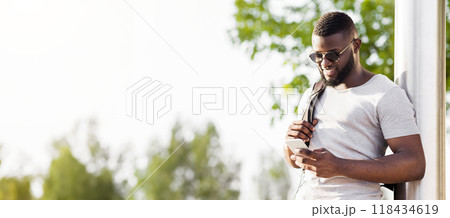 A man in sunglasses and a white t-shirt stands outdoors, using his smartphone. He has a backpack on and a slight smile on his face. A man in sunglasses and a white t-shirt stands outdoors, using his smartphone. He has a backpack on and a slight smile on his face. 118434619