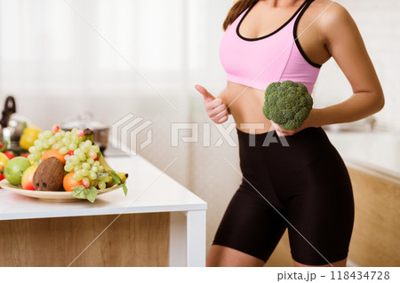 A woman wearing workout clothes holds a head of broccoli in her kitchen. A bowl of fruit is on the counter in the background. 118434728