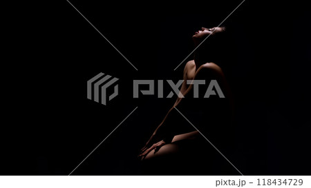 Naked young woman sitting in darkness with her arms on knee and looking up, panorama with copy space 118434729
