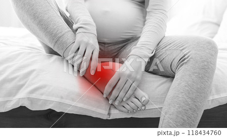 A pregnant woman is seated on a bed, massaging her lower leg, which is emphasized with a red highlight, suggesting discomfort or pain, black and white photo 118434760