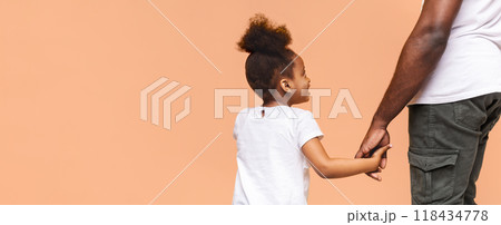 A little Black girl with a ponytail smiles as she holds her fathers hand. Her father is out of frame but his arm and hand are visible. The background is a light orange, likely studio wall, copy space A little Black girl with a ponytail smiles as she holds her fathers hand. Her father is out of frame but his arm and hand are visible. The background is a light orange, likely studio wall, copy space 118434778