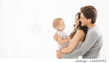 A young couple smiles as they hold their baby in their arms. They are standing in front of a white background, and the baby is looking off to the side, copy space A young couple smiles as they hold their baby in their arms. They are standing in front of a white background, and the baby is looking off to the side, copy space 118434791