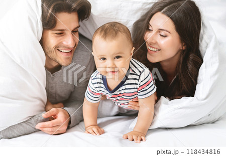 A happy family of three is playing under the covers in bed. The baby is crawling and looking at the camera while the parents are smiling and looking at their child. 118434816